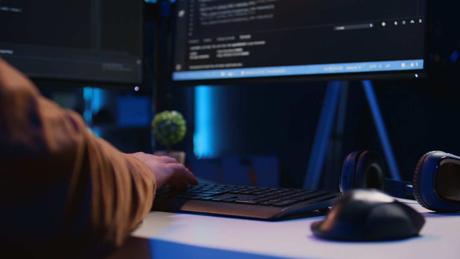 Iemand die op een toetsenbord zit te typen aan een bureau, met computerschermpjes waarop code te zien is, een koptelefoon en een muis ernaast.